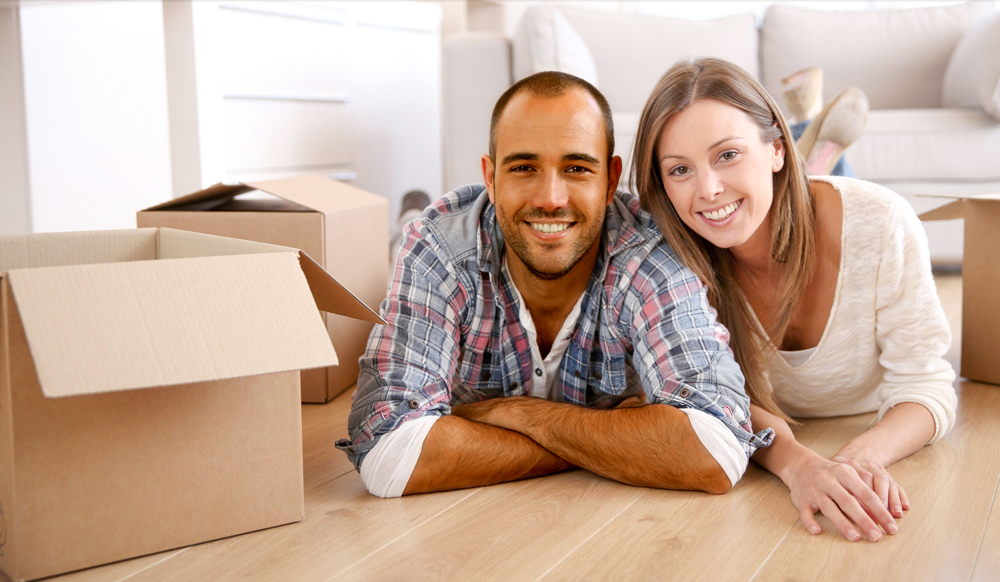 A cheerful man and woman lie on their stomachs on a wooden floor, facing the camera with wide smiles. They are surrounded by cardboard boxes in a well-lit living room, indicating a moving day.